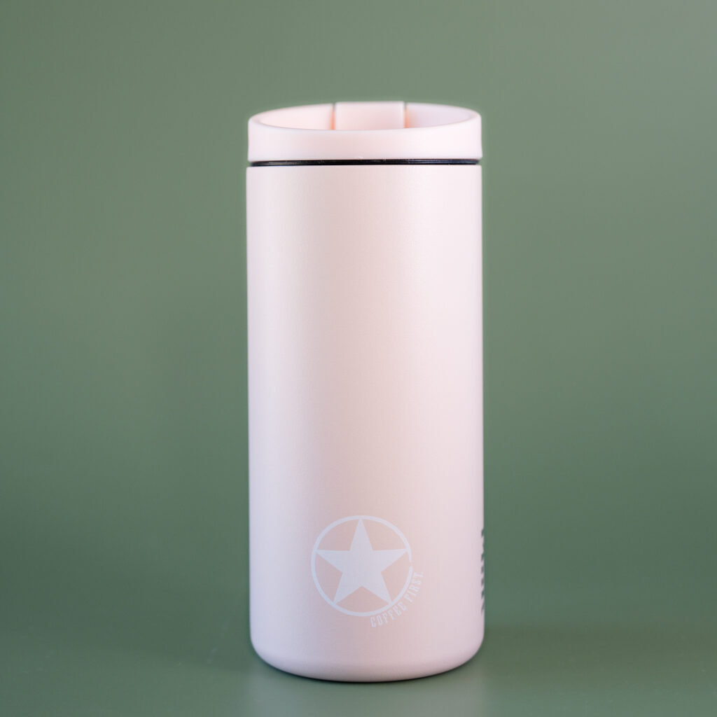 A pink miir flip tumbler with the rising star coffee logo that says Coffee first. The tumbler is set in front of a green background.