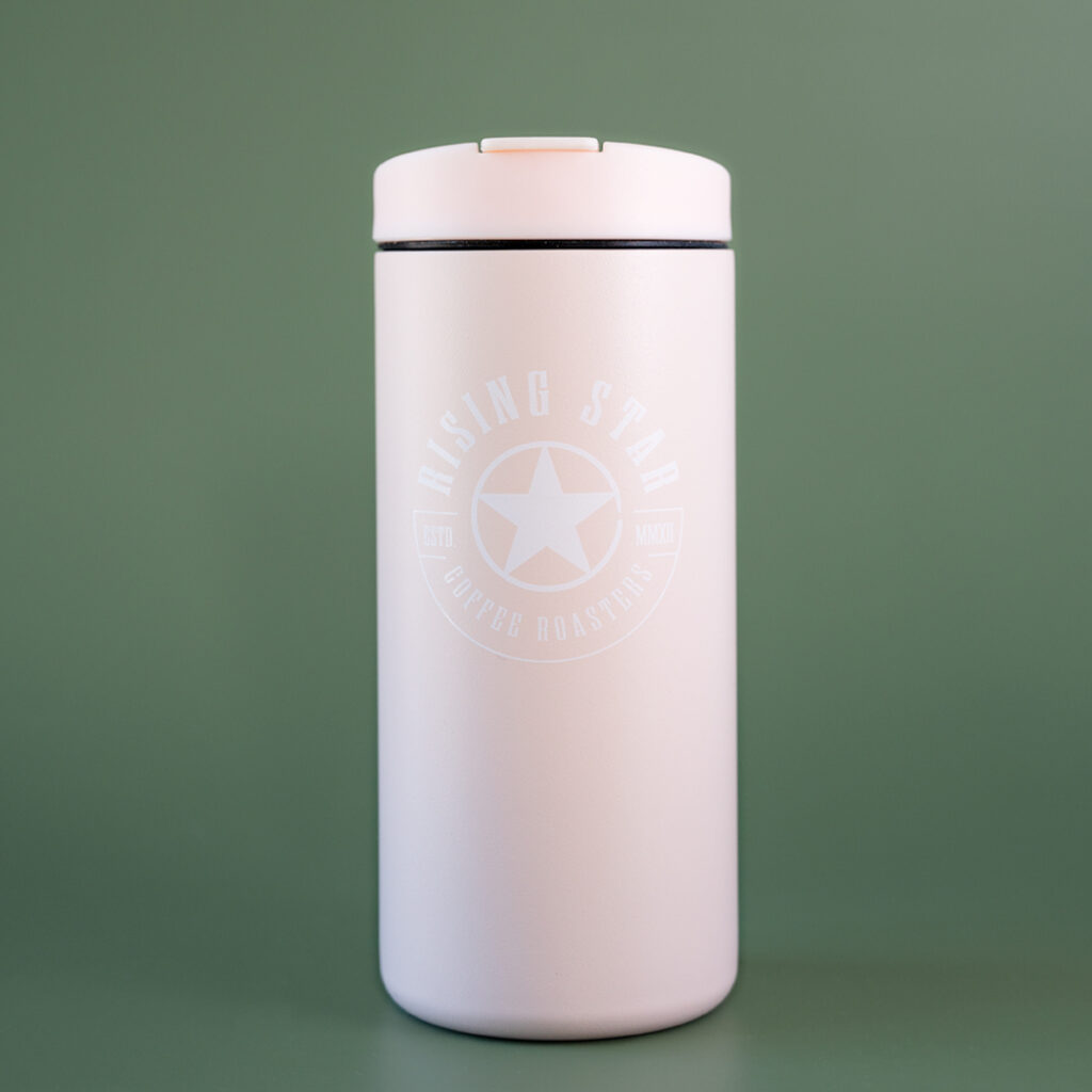 A pink miir flip tumbler with the rising star coffee seal on the front. The tumbler is set in front of a green background.