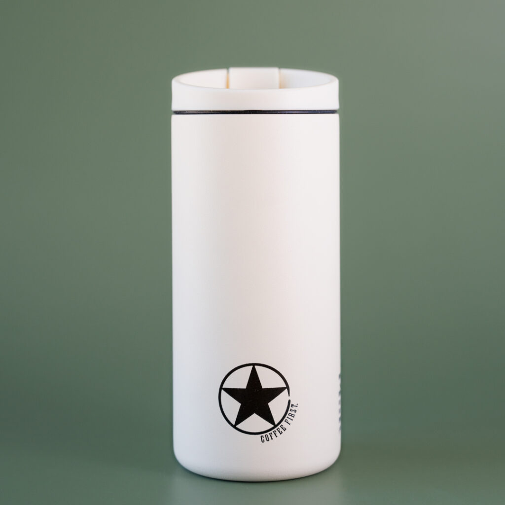 A sandstone white miir flip tumbler with the rising star logo with text that reads 'coffee first' on the bottom of the side of the cup. The tumbler is set in front of a green background.