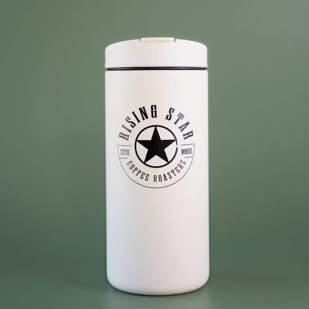 A sandstone white miir flip tumbler with the rising star coffee seal on the front. The tumbler is set in front of a green background.