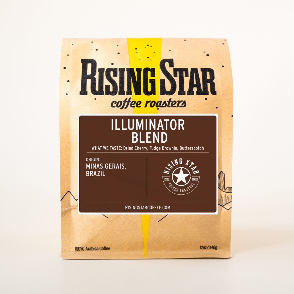 A 12oz bag of Rising Star Coffee Roasters' Illuminator Blend. The brown kraft paper bag features a yellow starbeam design and a dark brown label with white text. Flavor notes listed are dried cherry, fudge brownie, and butterscotch. The coffee’s origin is Minas Gerais, Brazil, and it is 100% Arabica.