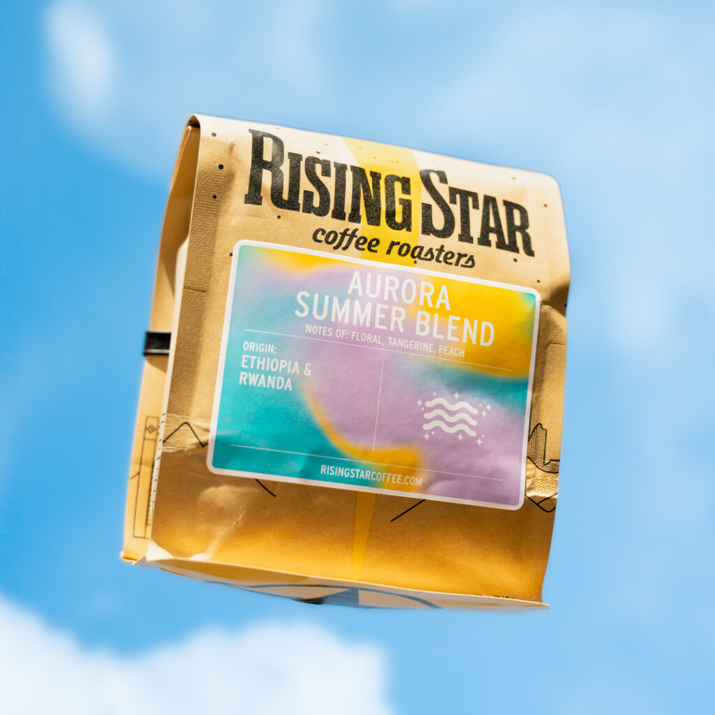A bag of Rising Star Coffee set on a bright blue sky background with fluffy clouds. The purple, teal, and golden yellow label reads - Aurora Summer Blend. Notes of Floral, Tangerine, Peach. Origin: Rwanda & Ethiopia. On the right side is a simplified line drawing of three white wiggles surrounded by white sparkles.
