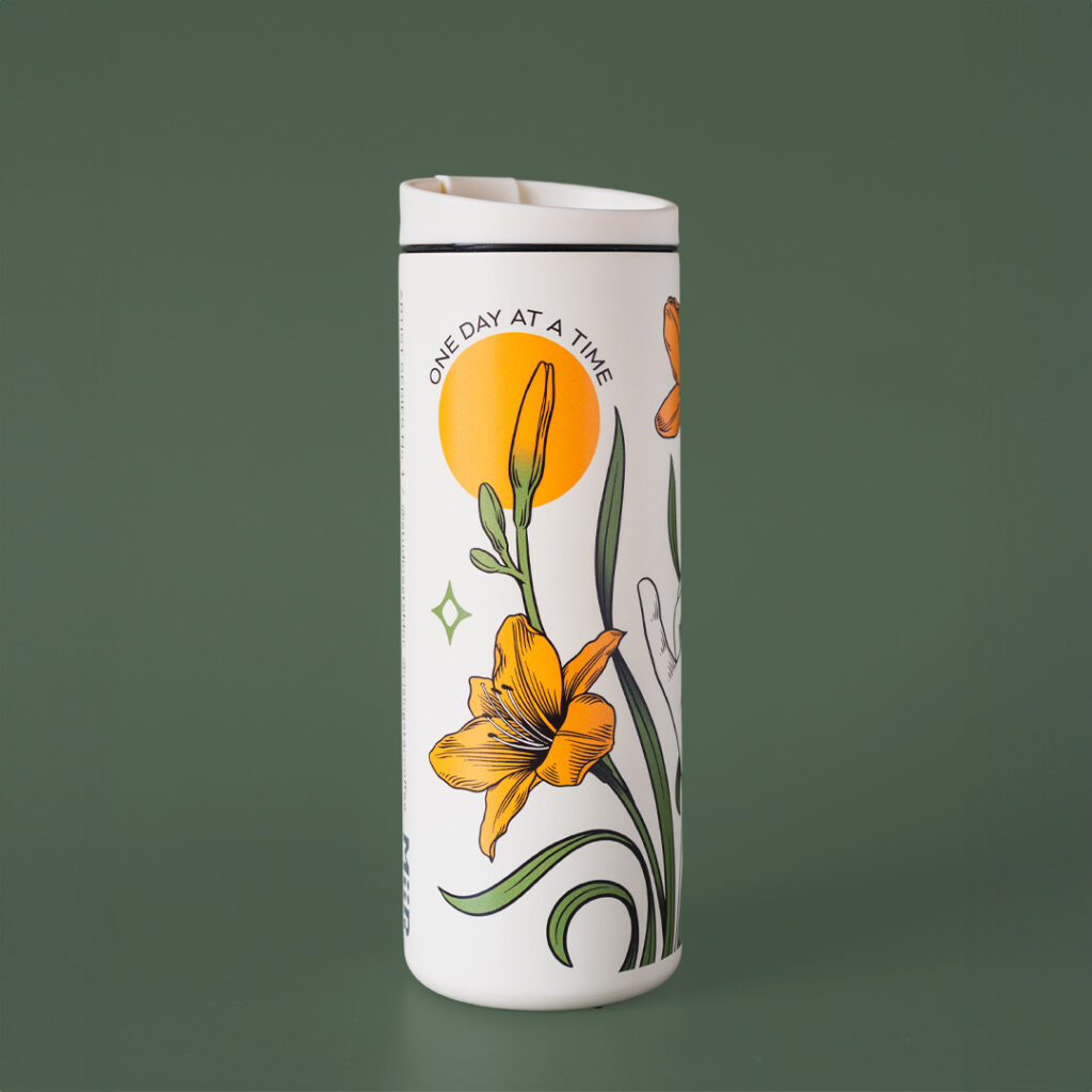 A white coffee travel tumbler with day lillies adorning the outside. It reads - 'one day at a time'