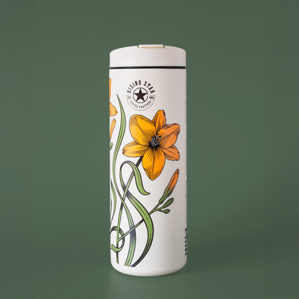 A white coffee travel tumbler with day lillies adorning the outside. It reads - 'one day at a time'