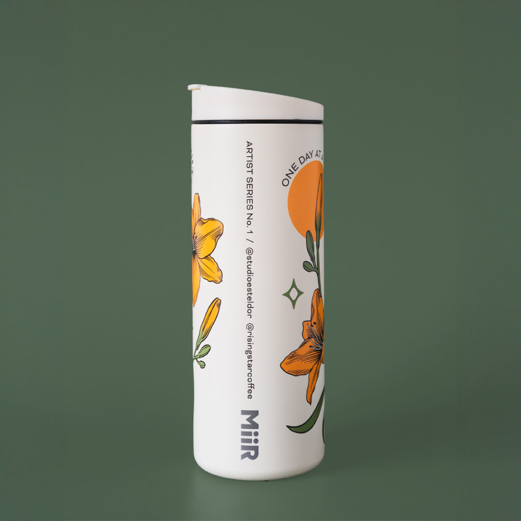 A white coffee travel tumbler with day lillies adorning the outside. It reads - 'one day at a time'