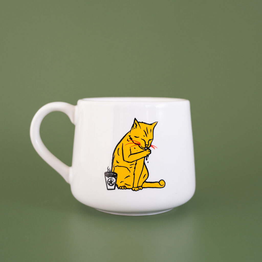 A white ceramic coffee cup that tapers towards the top. The artwork features a tabby cat licking their paw next to a cup of Rising Star Coffee