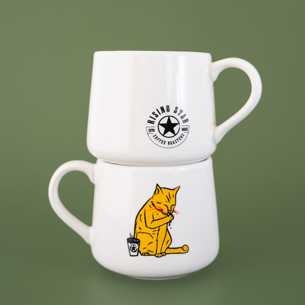 A white ceramic coffee cup that tapers towards the top. There is a nother of the same cup stacked on top that features the Rising Star Coffee logo in black. The artwork features a tabby cat licking their paw next to a cup of Rising Star Coffee