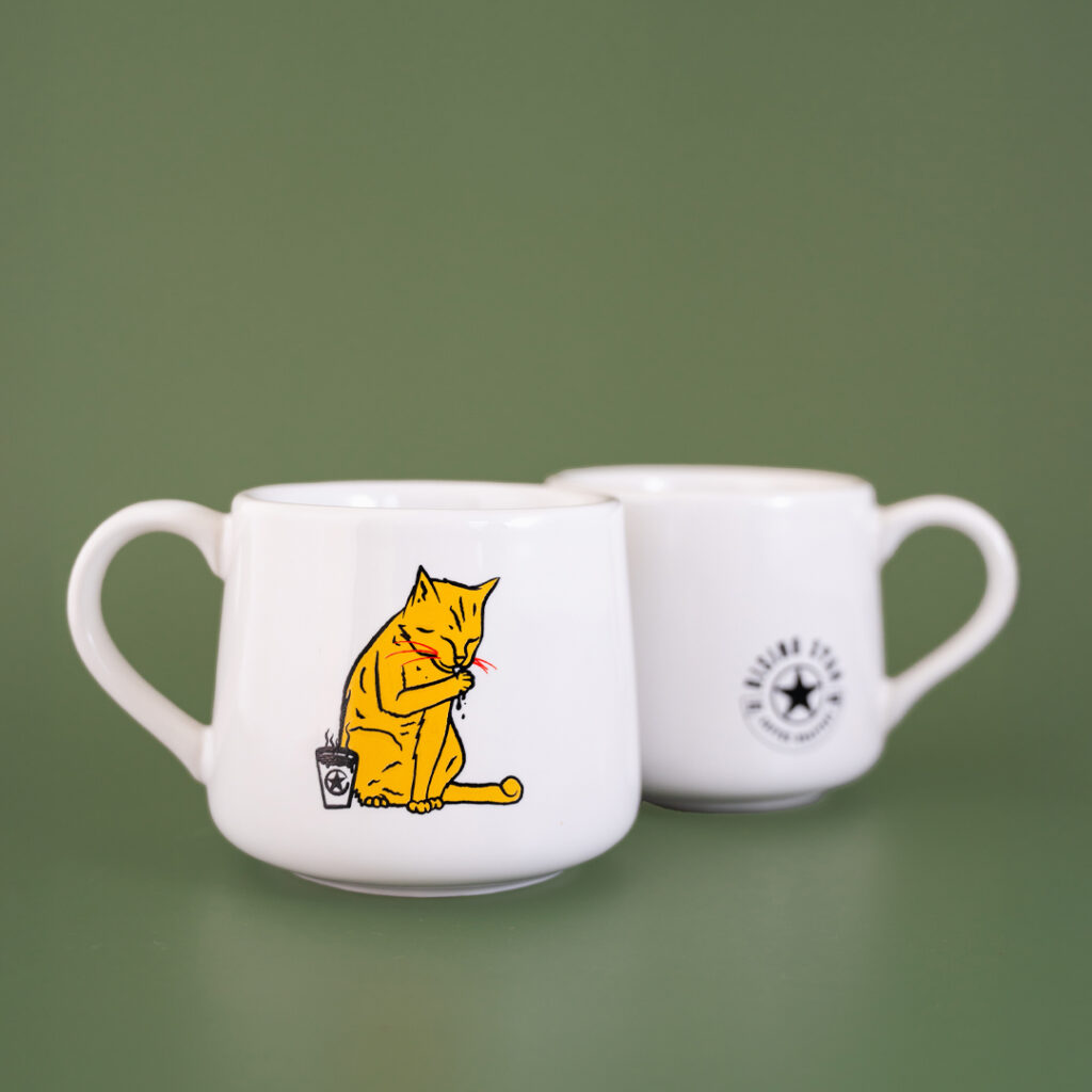 A white ceramic coffee cup that tapers towards the top. The artwork features a tabby cat licking their paw next to a cup of Rising Star Coffee There is the same cup behind this one turned the opposite way that features the Rising Star Coffee logo in black.