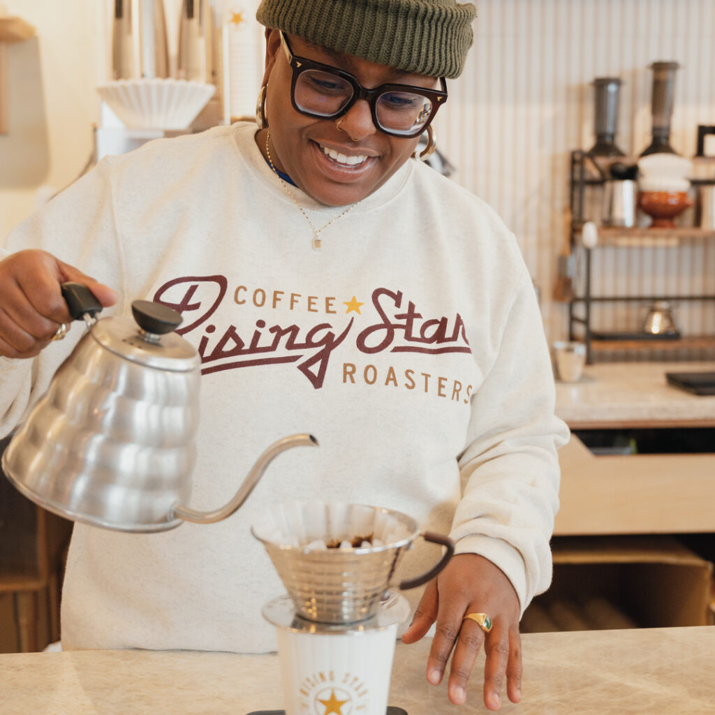 A photograph of a model wearing a tan crewneck sweatshirt that reads Rising Star Coffee Roasters. The model is brewing a pour-over.