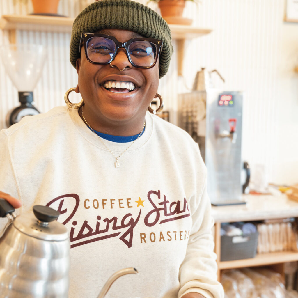 A photograph of a model wearing a tan crewneck sweatshirt that reads Rising Star Coffee Roasters. The model is brewing a pour-over.