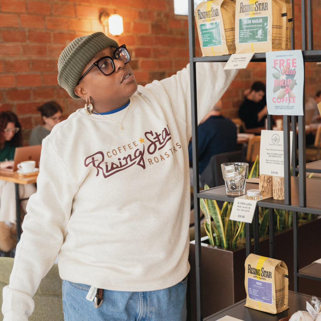 A photograph of a model wearing a tan crewneck sweatshirt that reads Rising Star Coffee Roasters. The model is stocking the display shelf