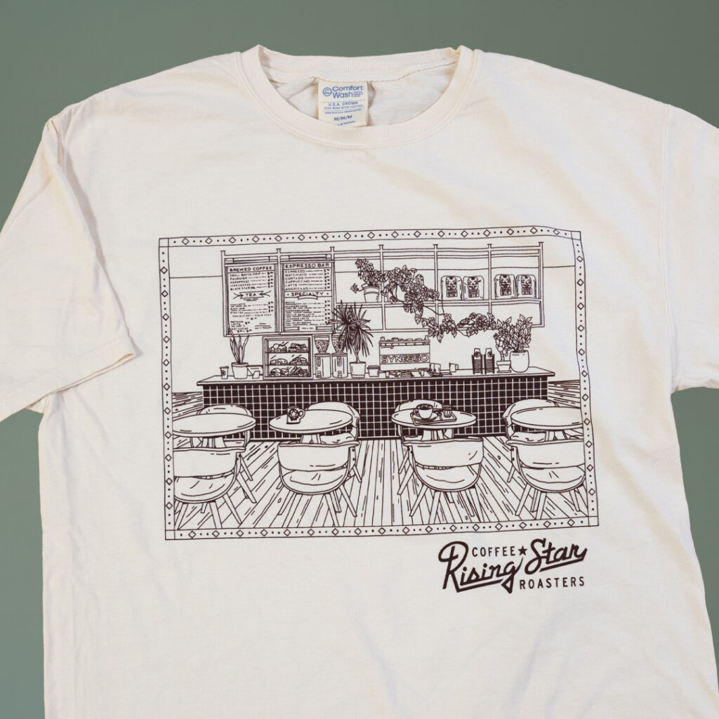 A cream colored t shirt with a line drawing of a cafe scene.