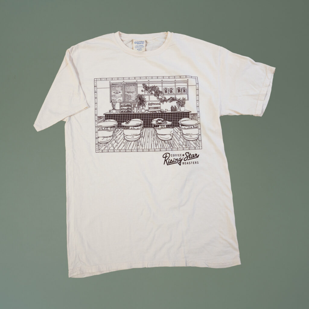 A cream colored t shirt with a cafe scene illustrated on it.