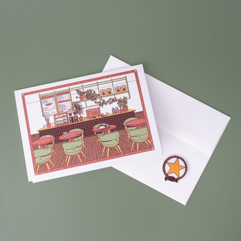 A photo of a greeting card with artwork of a cafe, an envelope and a sticker to seal said envelope.