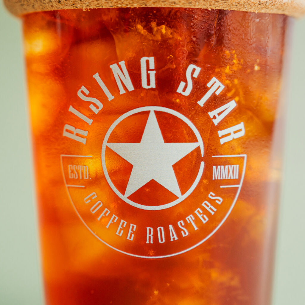 A close up of a glass iced drink tumbler full of iced coffee with a cork band and a sage green Rising Star Coffee logo on the side.