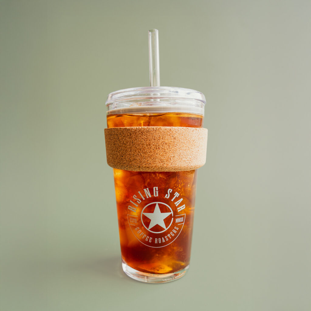 A glass iced drink tumbler full of iced coffee with a cork band and a sage green Rising Star Coffee logo on the side.