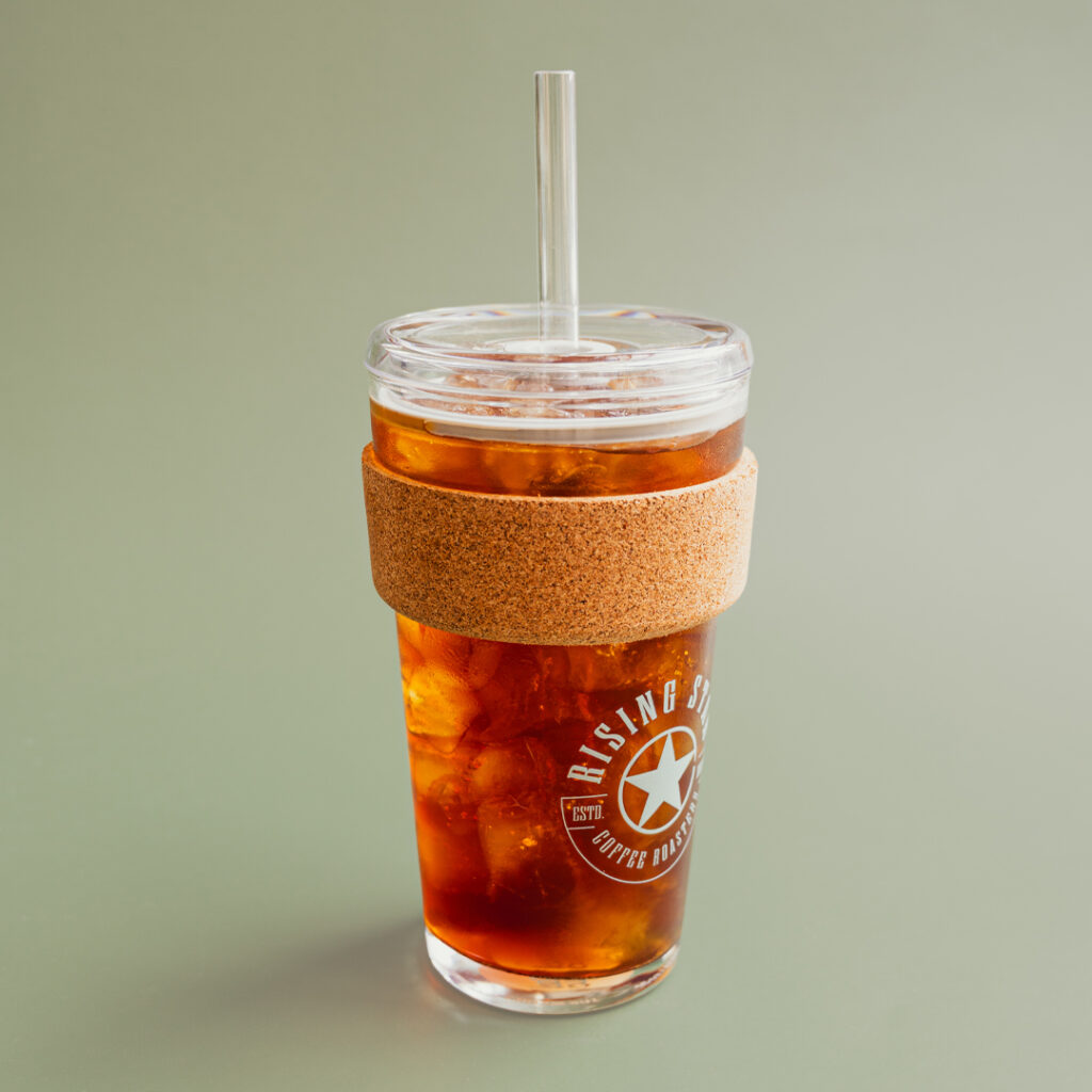 A glass iced drink tumbler full of iced coffee with a cork band and a sage green Rising Star Coffee logo on the side.