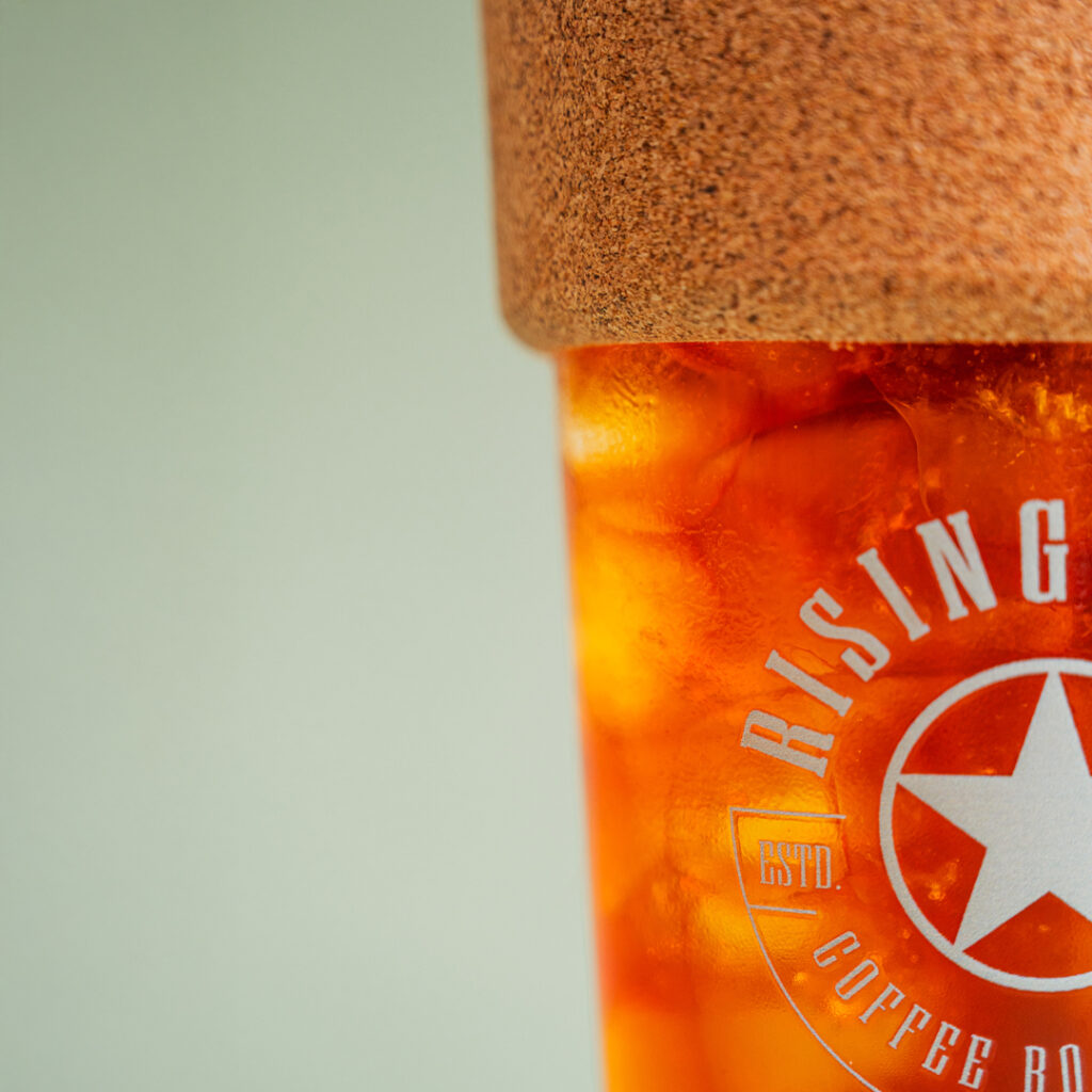 A close up of a glass iced drink tumbler full of iced coffee with a cork band and a sage green Rising Star Coffee logo on the side.