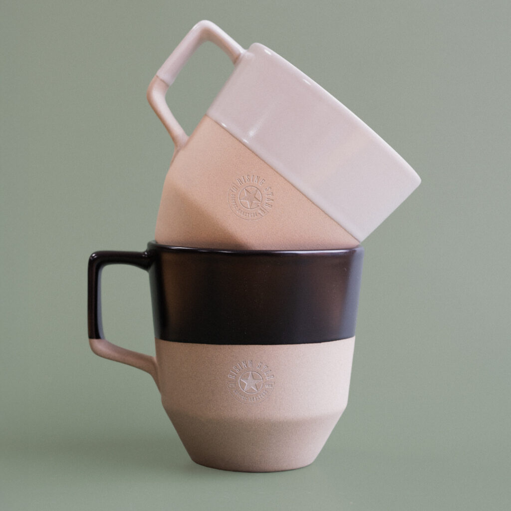 A black ceramic mug with a matching white one stacked off set on top. They have the Rising Star Coffee logo etched on the side.