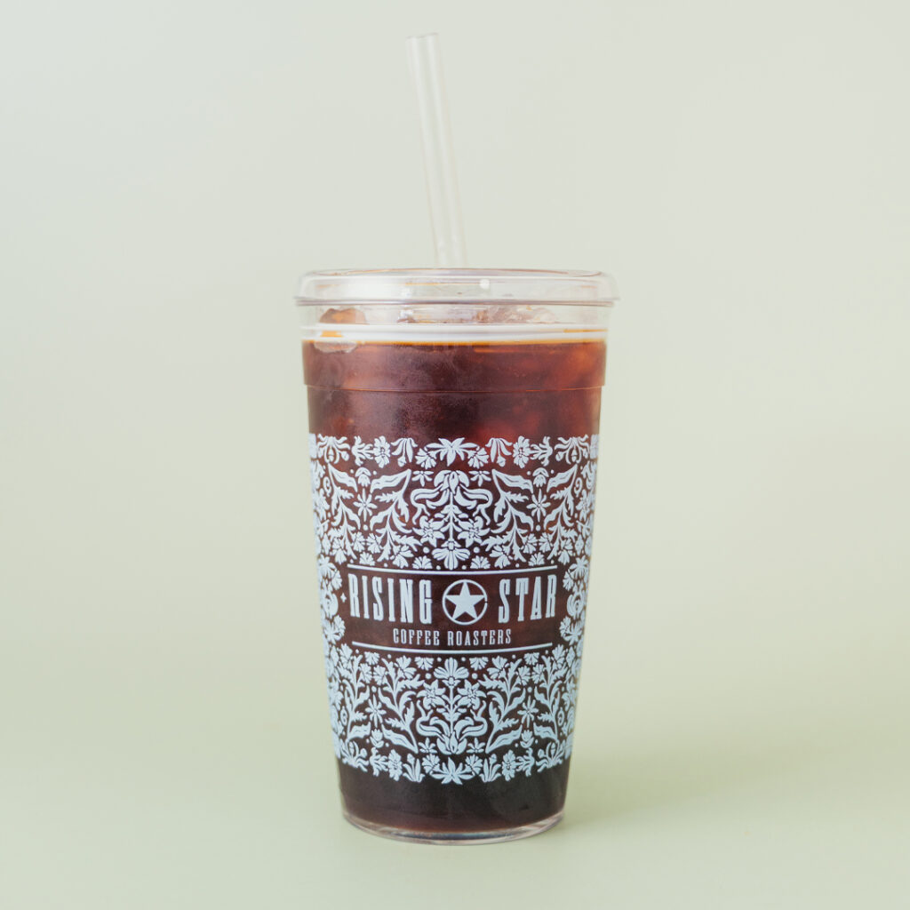 An iced coffee filled tumbler with an intricate floral design that wraps around the cup. The design is in a sky blue color that features the Rising Star Coffee Roasters logo.