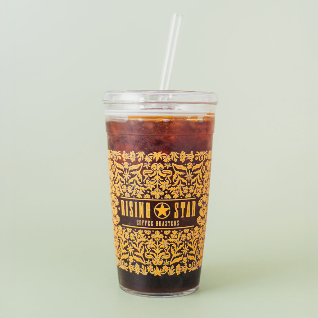 An iced coffee filled tumbler with an intricate floral design that wraps around the cup. The design is in a goldenrod color that features the Rising Star Coffee Roasters logo.