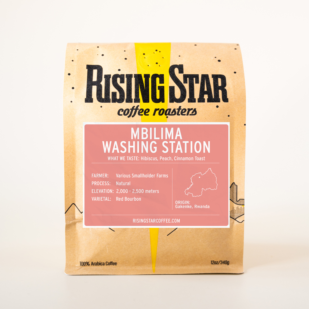 A tan bag of Rising Star Coffee with a light pink colored label and contrasting white text that reads: Mbilima Washing Station. Gakenke District, Rwanda. 100% Arabica coffee. What we taste: Hibiscus, Peach, Cinnamon Toast